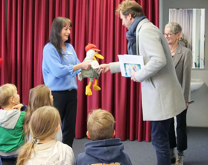 Frau mit Stofftier begrüßt den Bürgermeister, davor sitzen Kinder